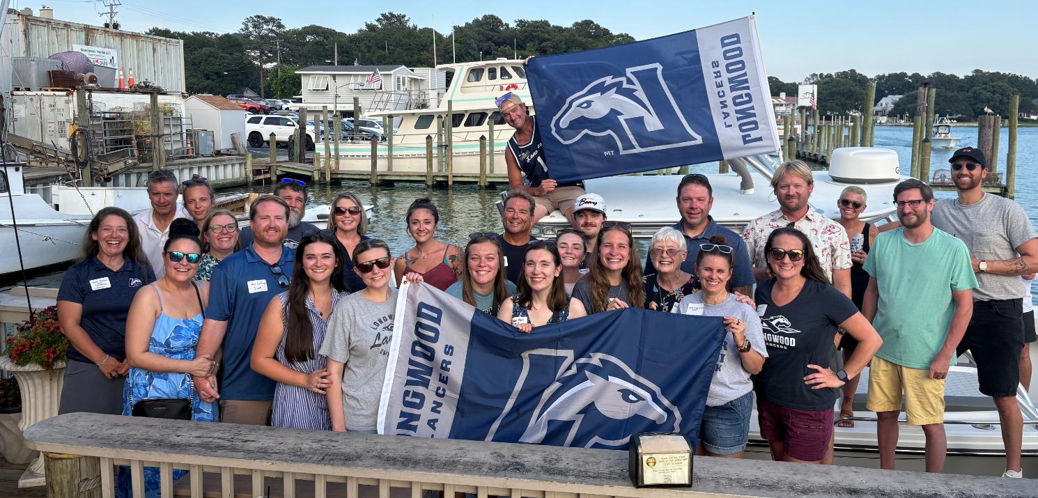 Longwood alumni gather at a regional event near the waterfront while holding Longwood Lancers flags.