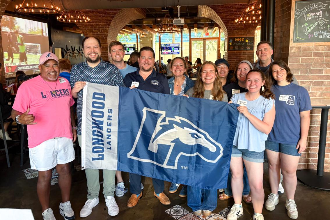 Longwood alumni and families gather at a NOVA regional event while holding a Longwood Lancers flag.