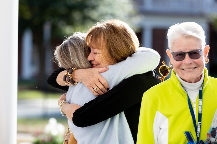 Two alumni share a warm hug outdoors during Homecoming 2025 while another woman in a bright Longwood jacket smiles nearby.