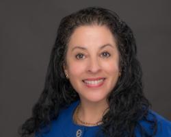 Professional headshot of a woman with long, curly black hair wearing a royal blue blazer and gold jewelry. She is smiling in front of a neutral gray background.