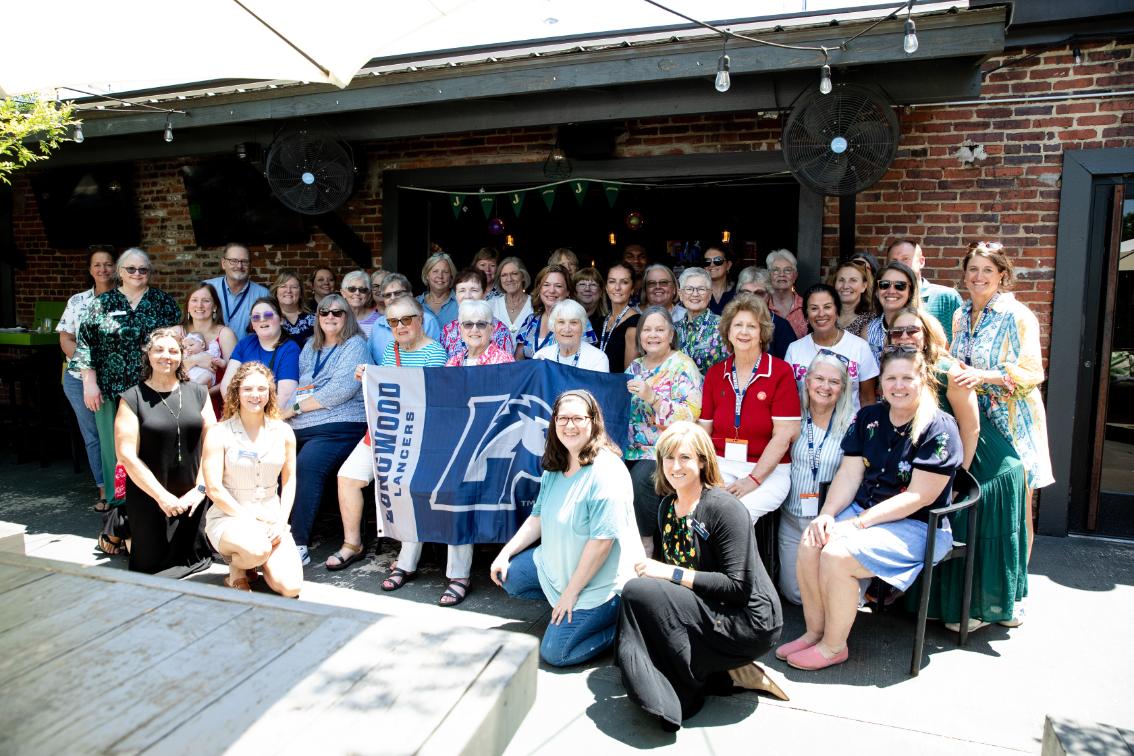 Longwood alumni and families gather at a regional event while holding a hometown Longwood Lancers flag.