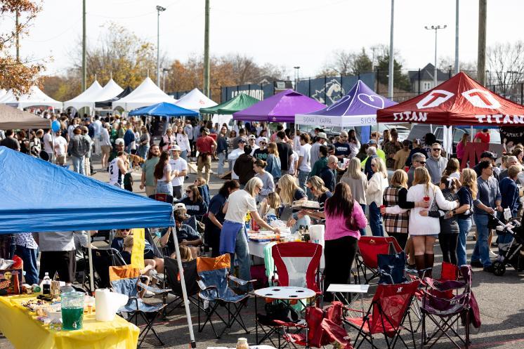 A wide view of a crowded Homecoming 2025 tailgate shows students, alumni, and families gathered among colorful tents, food tables, and lawn chairs in a lively outdoor setting.