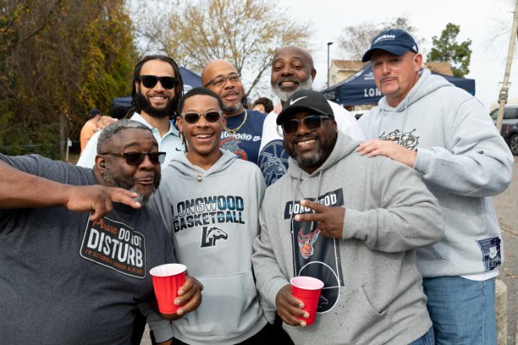 A group of alumni and fans smile and pose together at a Homecoming 2025 tailgate, wearing Longwood gear and holding drinks.