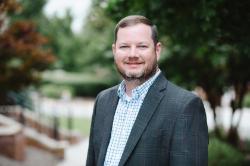 A man with short hair and a beard smiles outdoors, wearing a blue plaid jacket and checkered shirt, with greenery and a pathway in the background.