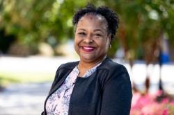 A woman smiles outdoors, wearing a dark jacket over a pink floral blouse, with bright sunlight and pink flowers in the background.