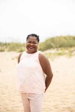 A smiling woman with short curly hair standing on a sandy beach with grassy dunes behind her. She is wearing a light pink sleeveless textured top and matching pants.