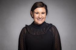 A woman with short brown hair in a side part, smiling in front of a light gray studio background. She is wearing a sheer black blouse with ruffles and subtle polka dot texture.