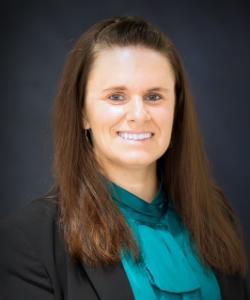 A woman with long straight brown hair smiling in front of a dark backdrop. She is wearing a dark blazer and a teal blouse with a pleated neckline.