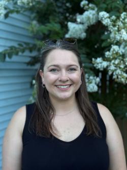 A woman with long brown hair and sunglasses resting on her head, smiling and standing in front of flowering bushes and light blue siding. She is wearing a sleeveless black top and hoop earrings.