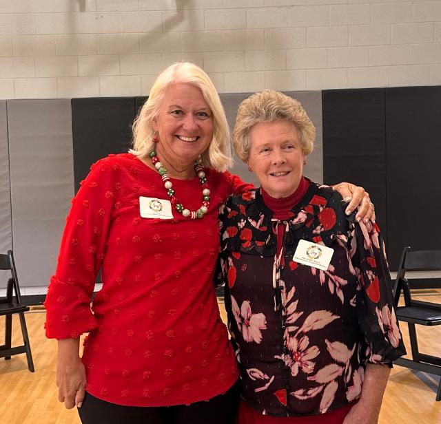 Two women are standing side by side, smiling. The woman on the left has blonde hair, wearing a red blouse and a festive necklace. The woman on the right has short, light hair, and is in a dark floral top with a red turtleneck. They both have name tags and are in a gym-like setting.