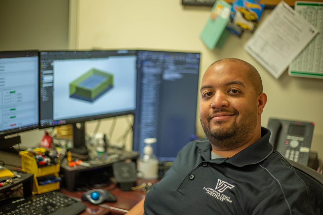 Courtesy of Virginia Tech Transportation Institute Man seated at a desk with dual monitors displaying design software, wearing a navy polo shirt embroidered with 