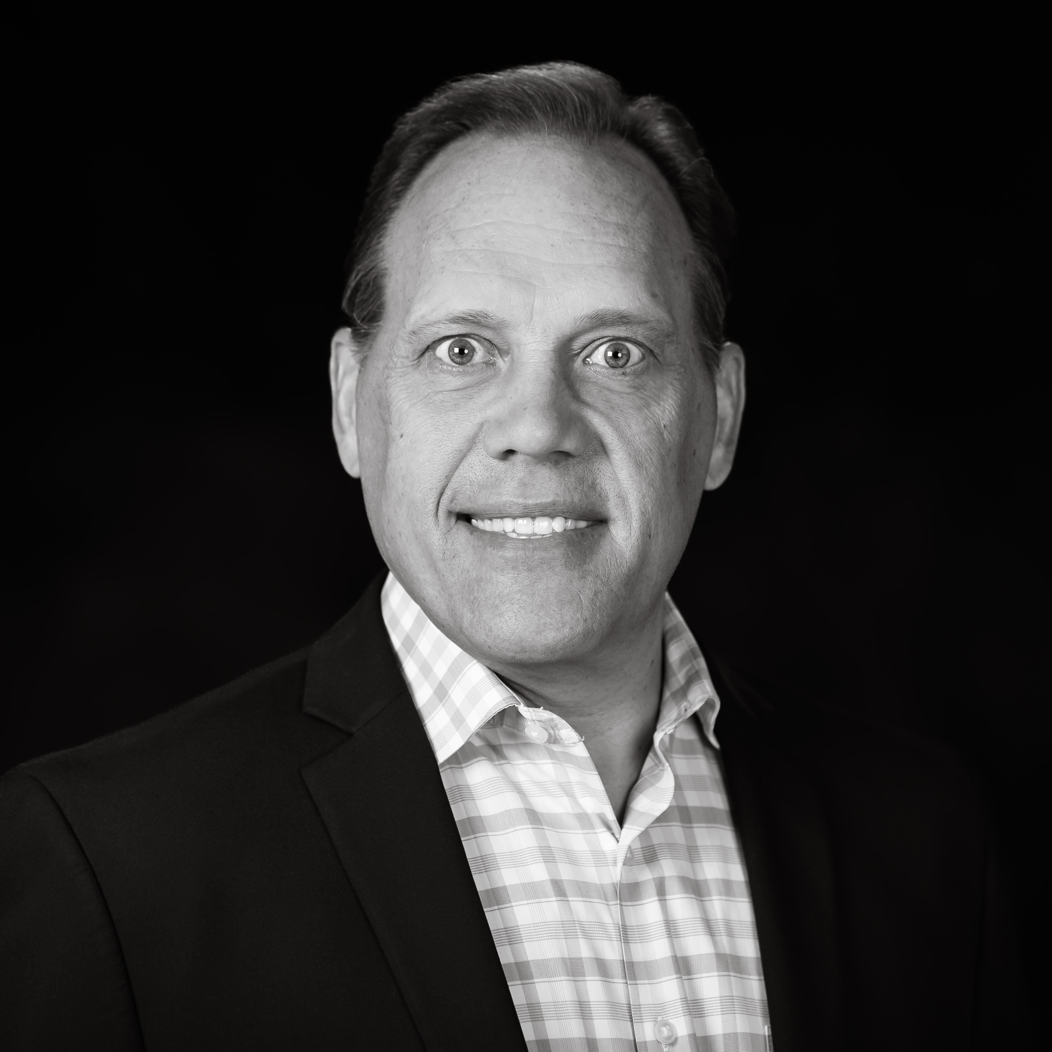 Black and white portrait of a man wearing a suit jacket over a checkered dress shirt, smiling at the camera with a dark background.