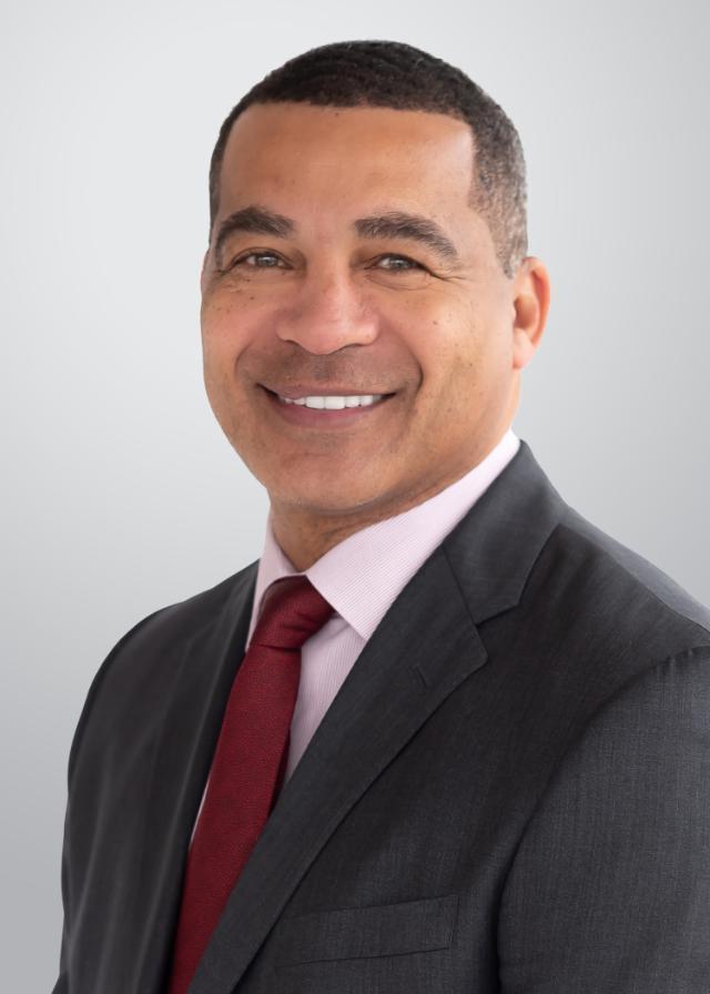 The image is a professional headshot of a middle-aged man with short black hair, a warm smile, and a confident expression. He is wearing a dark suit, a light pink dress shirt, and a red tie. The background is a neutral gray gradient.