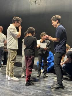 Students stand in a small circle onstage during a theater workshop or rehearsal activity.