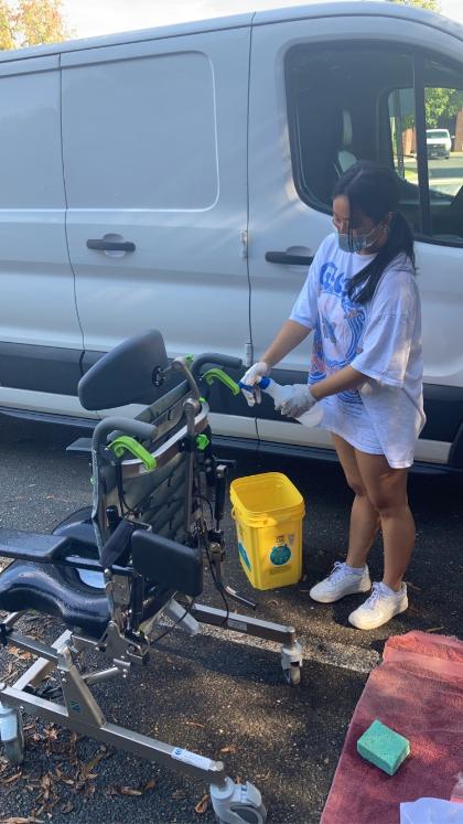 Olivia Outhong working during the Children’s Assistive Technology Wash A person wearing gloves and a mask uses a spray bottle to clean wheeled medical equipment outdoors beside a yellow bucket, with a white van parked in the background.