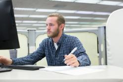 Student sits working on desktop computer - A person in a blue checkered shirt sits at a desk in an office, holding a pen and looking at a computer monitor, with a notebook, keyboard, and mouse on the desk and cubicles in the background.