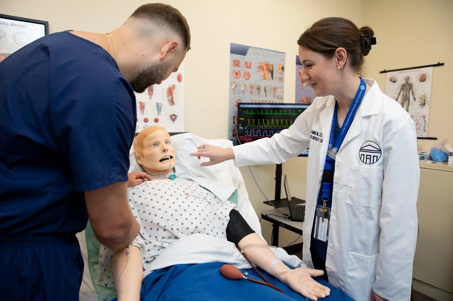 - Two individuals in a clinical setting work with a medical training mannequin on a hospital bed.