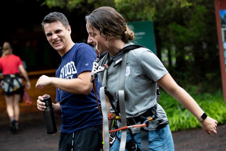 Two students laugh together at the ropes course. - Two students laugh together outdoors during a ropes course activity. One holds a water bottle while the other wears a climbing harness. They appear mid-conversation, smiling under overcast light. Trees and wooden structures form the casual, adventurous background.