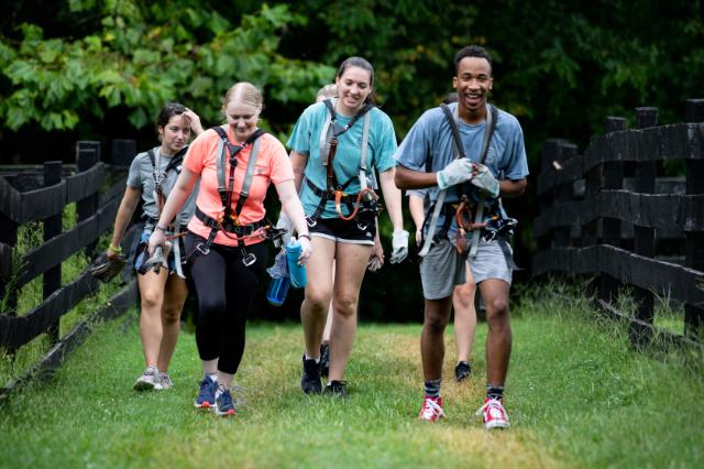 Students walk with harnesses to the ropes course during the 2021 leadership retreat - Four students wearing harnesses and gloves walk together along a grassy path bordered by a wooden fence. They smile and chat, appearing relaxed after an outdoor adventure activity. The group is dressed in athletic clothing and carrying water bottles. Lush green trees form a bright, natural backdrop.