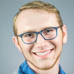 - Smiling person with short light hair and neatly trimmed beard wearing blue glasses. Dressed in a blue collared shirt under soft studio lighting. Face turned slightly toward the camera with a warm, friendly expression. Background is smooth gray, keeping the focus on the subject’s face.