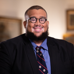 -Person smiling in a professional portrait. They are wearing glasses, a black suit jacket, a blue shirt, and a patterned tie. The background is softly lit with framed artwork on the walls. The setting appears to be an indoor office or academic environment.