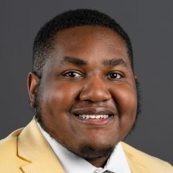 -Person smiling in a professional portrait. They are wearing a light yellow suit jacket, white shirt, and patterned tie. The background is a plain dark gray, creating a clean and formal look. The lighting evenly highlights the face and expression.
