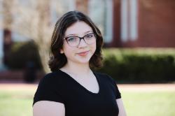 Carly Turner -Person smiling outdoors on a sunny day. Wearing glasses and a black V-neck shirt. Short brown hair styled in loose waves. Brick building and green lawn softly blurred in the background.