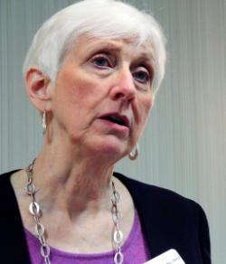 A woman with short white hair speaks indoors, wearing a purple top, black jacket, and a silver chain necklace.