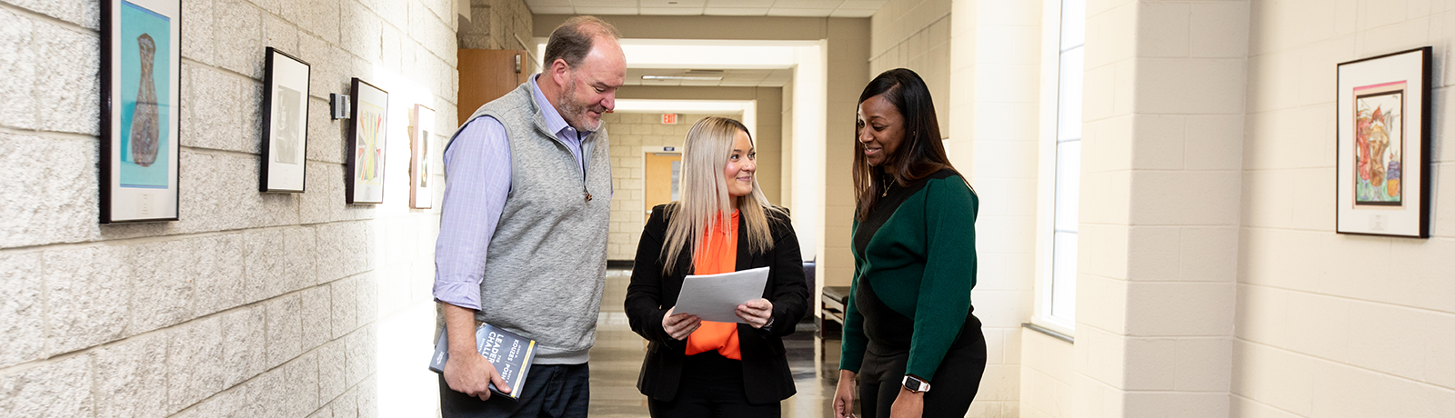 Three colleagues smile and talk while reviewing papers together in a bright hallway lined with framed artwork.