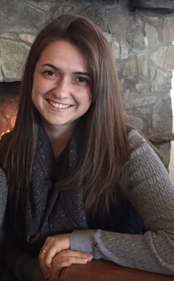 Jessica McCracken smiles while seated in front of a stone fireplace, wearing a gray sweater and dark scarf.