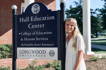 MaddieHommey stands next to the Hell Education Center sign. - A smiling woman in a white dress stands beside the Hull Education Center sign on Longwood’s campus. Sunlight highlights her long blonde hair as she poses next to the black and white sign. Brick walls, columns, and greenery frame the background. The scene feels bright and welcoming outdoors.