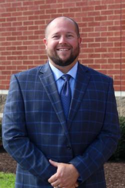 Smiling man with a short beard wearing a blue plaid suit jacket, light blue shirt and navy tie, standing in front of a red brick wall with his hands clasped.