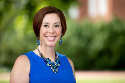 Sarah Tanner Anderson 2020 - A woman smiles while standing outdoors in front of soft green foliage. She wears a bright blue sleeveless top with matching blue and green statement jewelry. Her short brown hair is neatly styled, and she looks directly at the camera. The background is blurred, highlighting her as the focal point of the image.