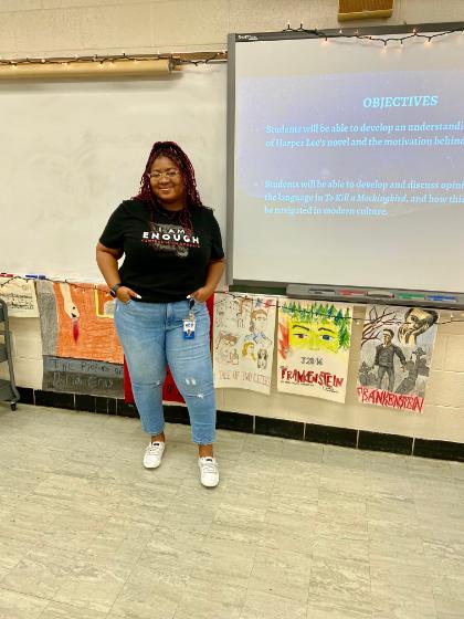 Sherae_Mcrae - A teacher stands smiling beside a classroom projector screen displaying lesson objectives. She wears a black “I Am Enough” T-shirt, jeans, and white sneakers. Colorful student artwork inspired by classic literature hangs along the wall behind her. The classroom setting includes a whiteboard, string lights, and a tiled floor.