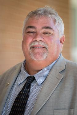 Vertical photo of Dr. Danny Grounard Visiting Assistant Professor Coordinator Educational Leadership Endorsement - A man with short gray hair and a neatly trimmed goatee smiles at the camera. He is wearing a light gray suit jacket, a striped dress shirt, and a patterned dark tie. Soft, warm lighting highlights his face against a blurred tan background. His expression is friendly and professional.