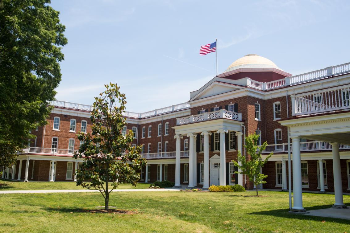 Rotunda - A stately red-brick campus building features white columns and a domed roof. An American flag waves above the central portico. Manicured lawns and young trees frame the front courtyard. Bright summer light highlights the building’s classical architecture.