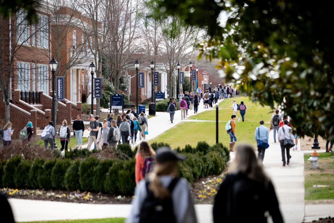 Students walking on Brock Commons between classes - Students walk along a busy campus sidewalk lined with brick buildings and lamppost banners. Groups move in both directions, many carrying backpacks between classes. Early spring trees and landscaped bushes frame the walkway on both sides. The scene captures an active, energetic moment in the heart of campus.