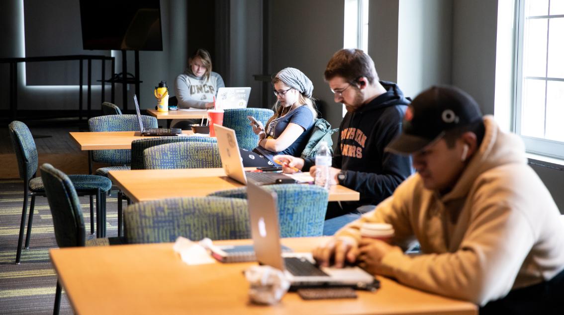 Students working at their laptops in the student union - Students sit spaced out at tables in a quiet campus study area. Several work on laptops while others take notes or check their phones. Natural light from large windows brightens the room’s muted tones. The scene reflects focused, individual study in a relaxed environment.