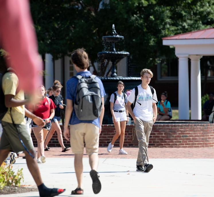 Students walking around the Rotunda fountain - Students walk across a sunny campus courtyard between classes. A tiered fountain sprays water in the background. Several students carry backpacks while others sit along a brick wall. Trees and white-columned walkways frame the busy outdoor scene.