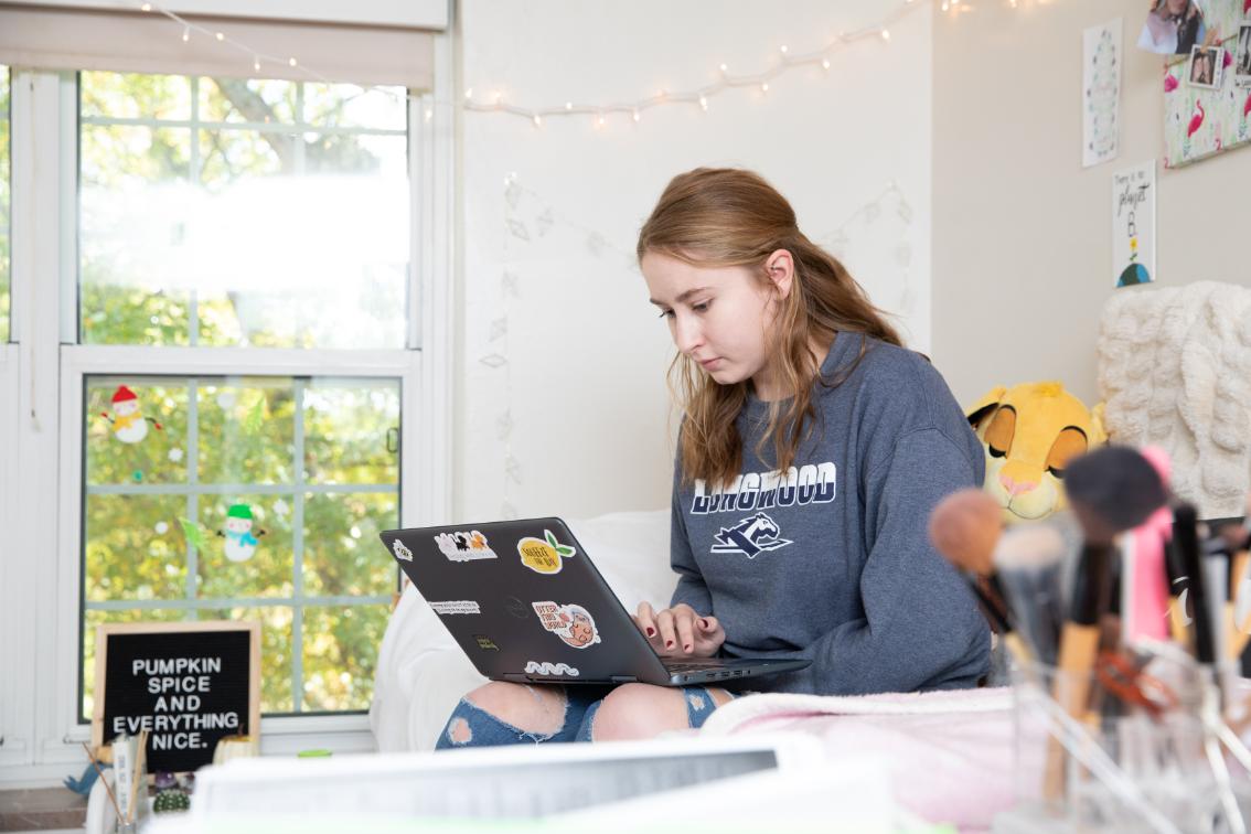 Student working on a laptop sitting on the bed in her residence - A student sits on a dorm room bed working on a laptop. Soft string lights and wall décor hang behind her. A window with seasonal stickers brings in bright natural light. Makeup brushes and a small sign sit in the foreground. hall room
