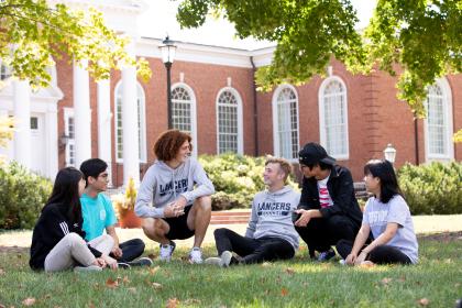 Students sitting on the grass talking outside Eason Hall - A group of six students sit together in the grass on a sunny day. They are talking and laughing beneath leafy green trees. Red-brick academic buildings with tall white columns rise behind them. The scene feels relaxed and welcoming, capturing campus life outdoors.