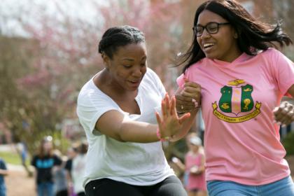 Two students run together.  - Two students laugh and move together during an outdoor campus event. One wears a pink Alpha Kappa Alpha shirt while the other wears a white top. They appear mid-step with expressive gestures and smiles. Other students and flowering trees blur in the background.