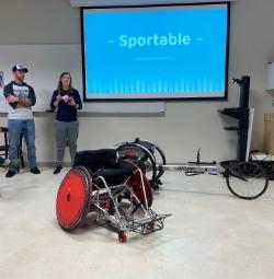 Sportable Organization - An adaptive sports wheelchair and handcycle are displayed in a classroom. Two presenters stand at the front of the room addressing an audience.