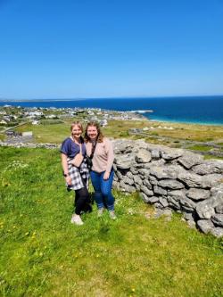 Two TR students pose at a cliff in Ireland - Two students stand together on a grassy hillside overlooking a coastal village. A low stone wall runs beside them in the foreground. The blue ocean stretches behind the town under a clear sky. The scene captures a scenic cliffside view along the Irish coast.
