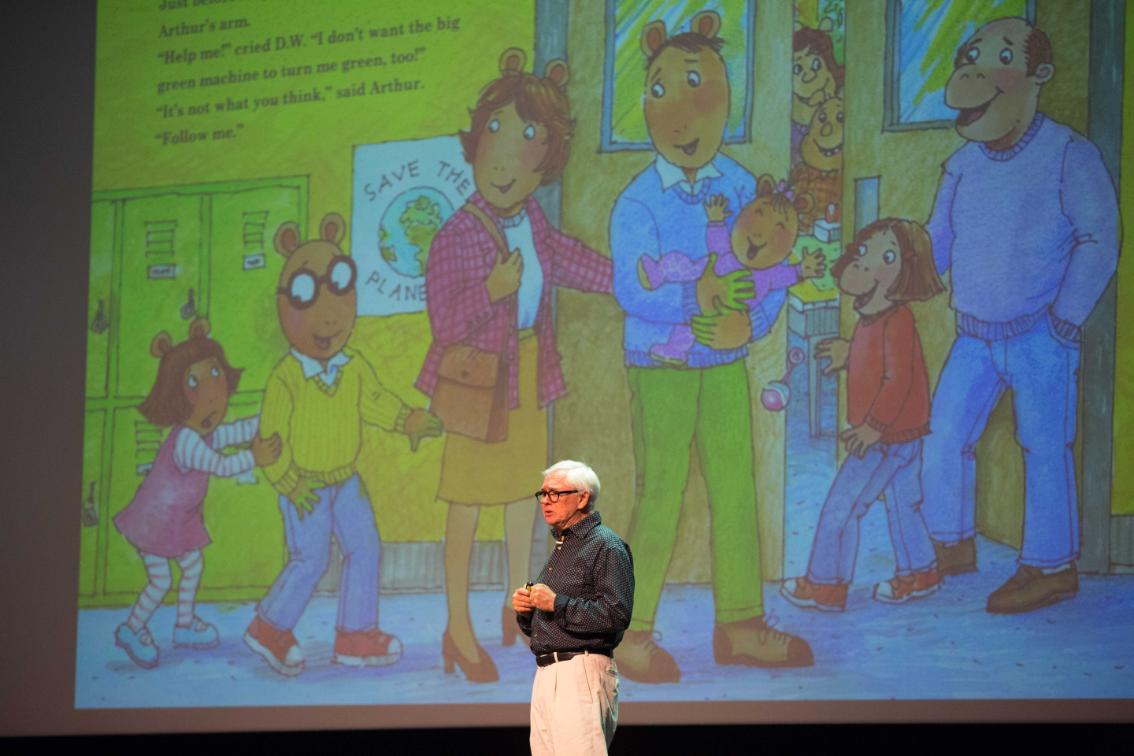Marc Brown at the Virginia Childrens Book Festival - A speaker stands onstage in front of a large projected illustration. The projection shows characters from the children’s book series Arthur. The presenter holds a microphone while addressing the audience. Colorful storybook art fills the screen behind him.