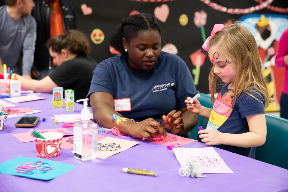 LCVA Valentines Family Workshop - Longwood students and community members work together at a Valentine’s-themed craft table. A woman in a Longwood Global Learning shirt helps a child with a pink paper craft. Art supplies, markers, and decorations are spread across a purple tablecloth. A decorated backdrop with hearts and flowers fills the room behind them.