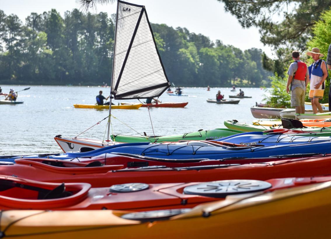 Kayaking on Wilck's Lake - Colorful kayaks are lined up along the shore of a lake. A small sailboat sits among the boats in the foreground. Paddlers in kayaks are spread out across the water in the distance. Two people stand on the bank near the launch area on a sunny day.