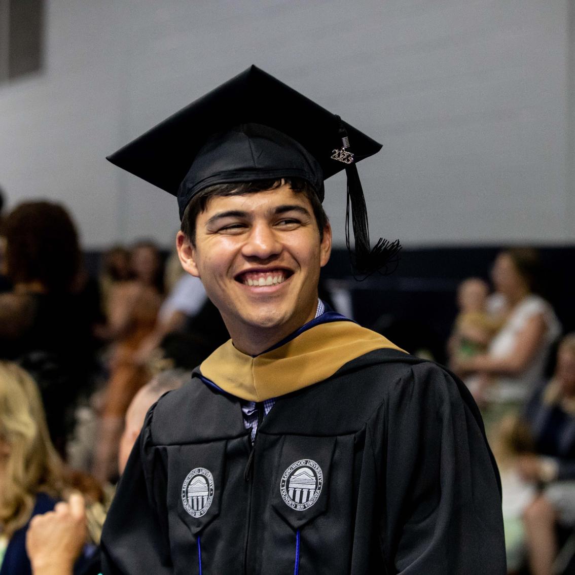Student during graduate commencement ceremony - A smiling graduate wears a black cap and gown with a tassel marked “2023.” The graduate looks to the side during a commencement ceremony indoors. A gold hood and Longwood University seal patches are visible on the gown. The background is softly blurred with other attendees in the venue.