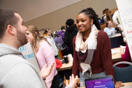 A student smiles while speaking with another student at a campus involvement fair. They stand near display boards and tables filled with information. The event is busy, with several students interacting in the background. A laptop on the table displays the handle “@afroh.longwood.”