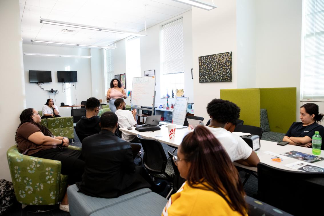 Student meeting in the Cookie Scott Lounge - Student leads a group discussion while standing near a whiteboard. Other students sit around tables and lounge seating, listening and participating. Laptops, drinks, and notes are visible across the workspace. The meeting takes place in a bright, modern campus lounge.